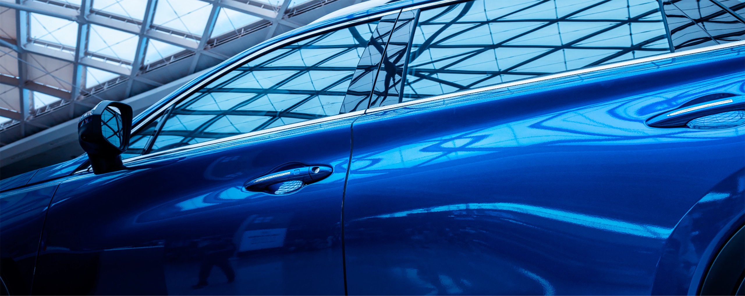 Blaues Auto seitlich in einem modernen, verglasten Gebäude, reflektiert Gittermuster der Decke auf der Karosserie.