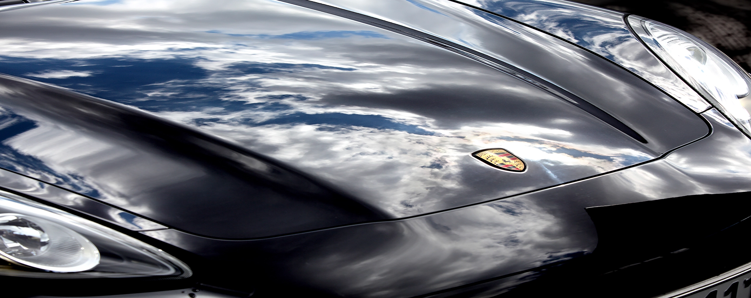 Reflektionen von Wolken auf der glänzenden Motorhaube eines schwarzen Autos mit erkennbarem Emblem.